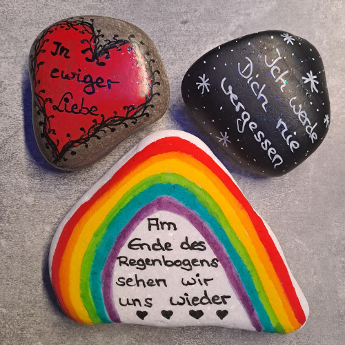 Trauer und Abschied, dargestellt durch drei Gedenksteine, ein Regenbogenstein mit Spruch, ein Stein mit einem Herz und Spruh und ein schwarzer Stein mit Spruch