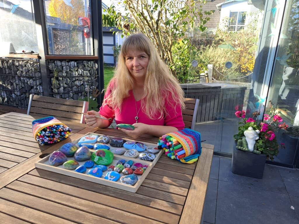 Auf dem Foto sitze ich am Tisch mit einem Tablett voller bemalter Steine, die ich gerade bemalt und gestaltet habe