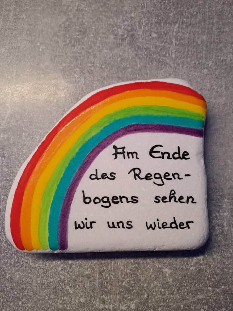 Am Ende des Regenbogens Gedenkstein ist ein weißer halbrunder Marmorstein, auf dem entlang der linken Seite ein großer bunter Regenbogen gemalt wurde und darunter steht Am Ende des Regenbogens sehen wir uns wieder
