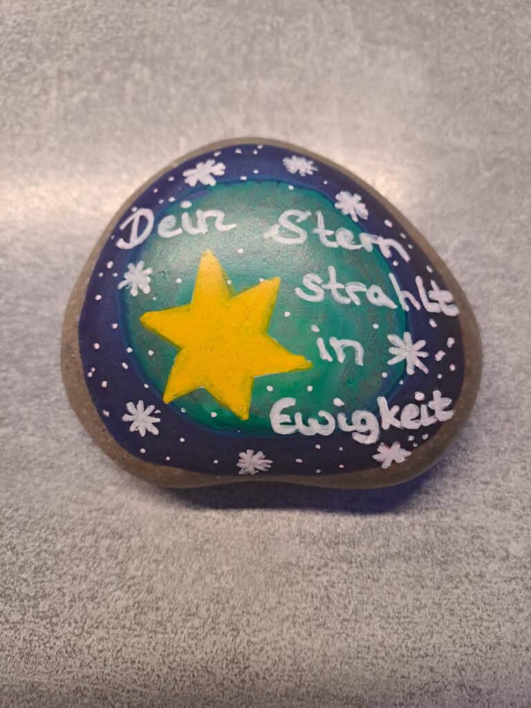 Dein Stern Gedenkstein Sternenkind, Dunkelblau-türkisfarbener Stein mit einem großen gelben Stern und der Aufschrift in weiß-rosa "Dein Stern strahlt in Ewigkeit" und kleinen weißen Sternen rundherum