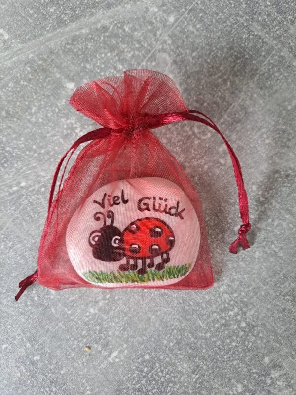 Glücksstein „Viel Glück“ für die Hosentasche mit Glückskäfer, weißer Stein mit rotem Marienkäfer und der Aufschrift Viel Glück, verpackt in einem roten Organzabeutelchen
