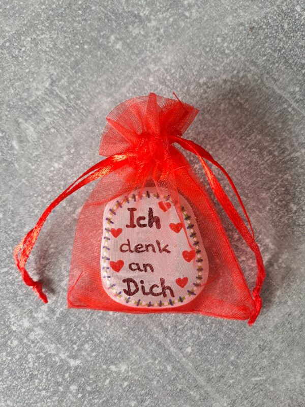 Ich denk an Dich Mutmacher für die Hosentasche, weißer Stein mit mintgrünem Hintergrund, Aufschrift ich denk an dich, kleinen Herzen und einer Bordüre bestehend aus verschiedenfarbigen kurzen Strichen und kleinen Punkten dazwischen, verpackt in ein rotes Organzasäckchen