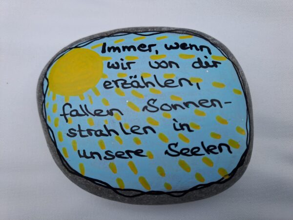 Sonnenstrahlen Spruch Stein, Hellgrauer Stein bemalt mit blauem Himmel und einer Sonne mit ganz vielen Strahlen und dem Spruch Immer, wenn wir von dir erzählen, fallen Sonnenstrahlen in unsere Seelen