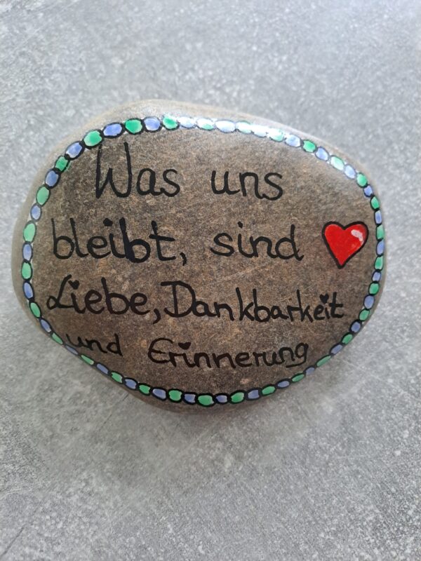 Was uns bleibt sind Liebe Dankbarkeit und Erinnerung Spruch Stein, mit schwarz auf einen grauen Stein geschrieben, verziert mit einem roten Herz und ringsherum mit einer Bordüre aus blauen und grünen großen Punkten im Wechsel