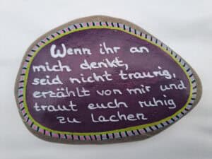 Wenn ihr an mich denkt Spruch Stein, hellgrauer Stein bemalt mit Lila Hintergrund und weißem Spruch, außen herum befindet sich eine mehrfarbige Bordüre