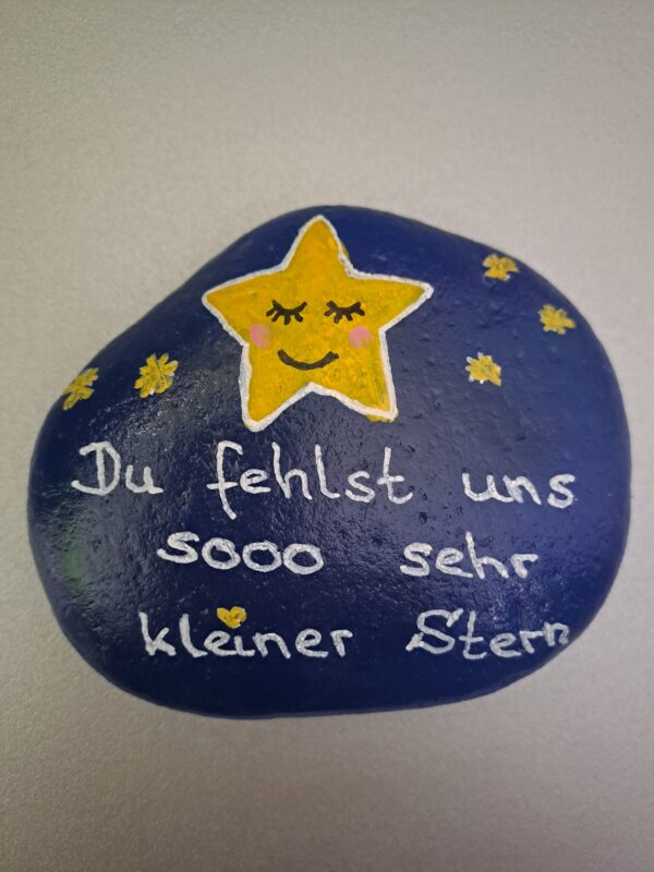 Du fehlst uns kleiner Stern, dunkelblauer Stein mit großem gelbem Stern und mehreren kleinen Sternchen und der weißen Aufschrift "Du fehlst und sooo sehr kleiner Stern"