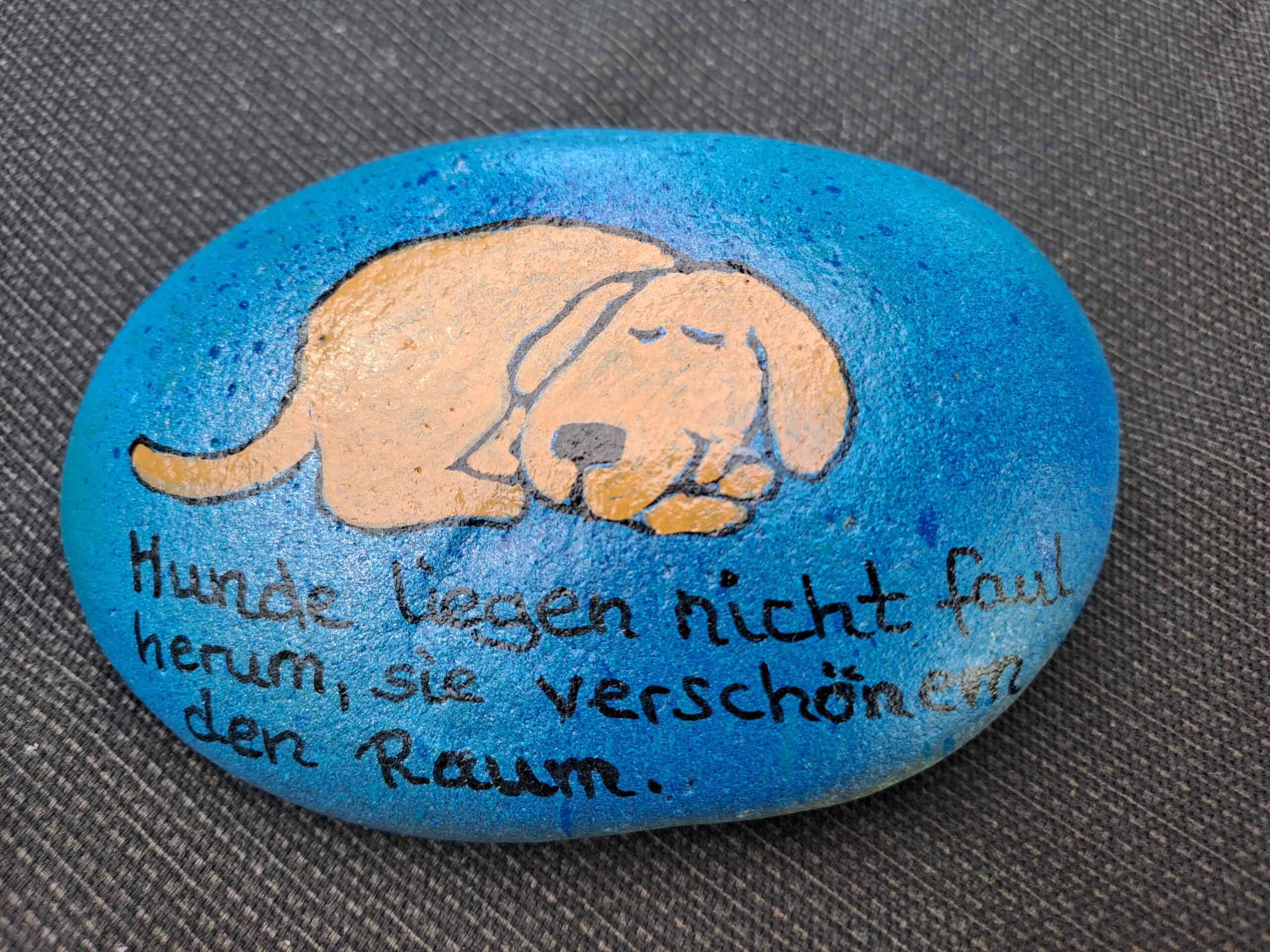 blauer Stein mit beigebraunem schlafenden Hund und dem Spruch "Hunde liegen nicht faul herum, sie verschönern den Raum"