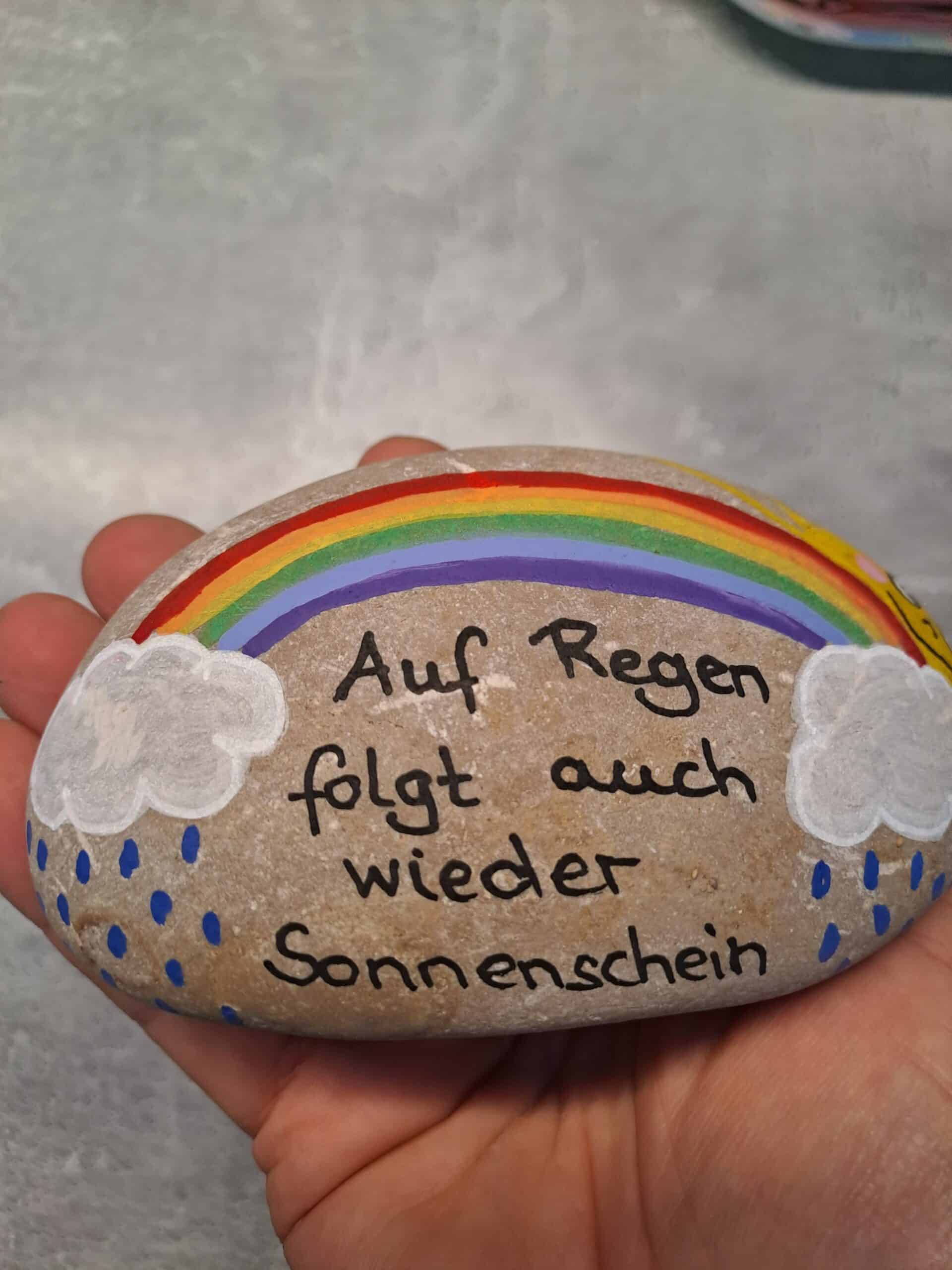 Stein mit buntem Regenbogen, zwei weißen Regenwolken, einer lachenden Sonne und dem Spruch "Auf Regen folgt auch wieder Sonnenschein"
