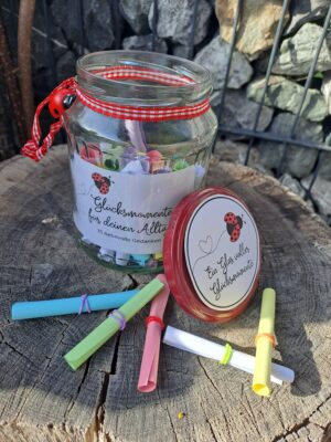 DIY-Sprücheset „Glücksmomente für deinen Alltag“ fertig in einem leeren Konservenglas, schön dekoriert mit einem rot-weißen Band, Etiketten auf Glas und Deckel sowie gefüllt mit Sprüchen, ein paar liegen daneben