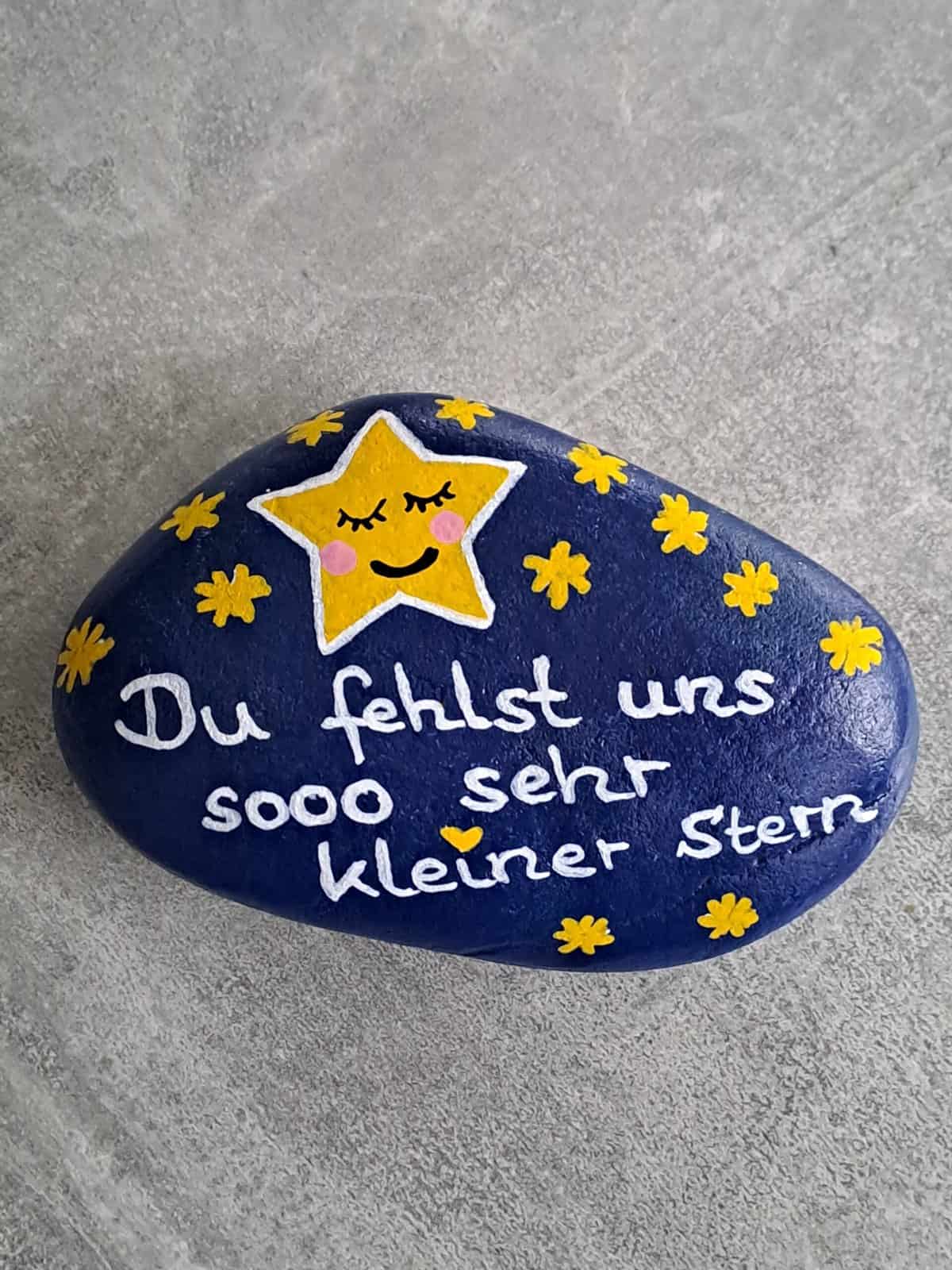 Dunkelblau lackierter Stein mit einem großen gelben Stern mit schlafendem Gesicht darauf, daneben viele kleine gelbe Steine und der Spruch Du fehlst uns sooo sehr kleiner Stern, Variante 1
