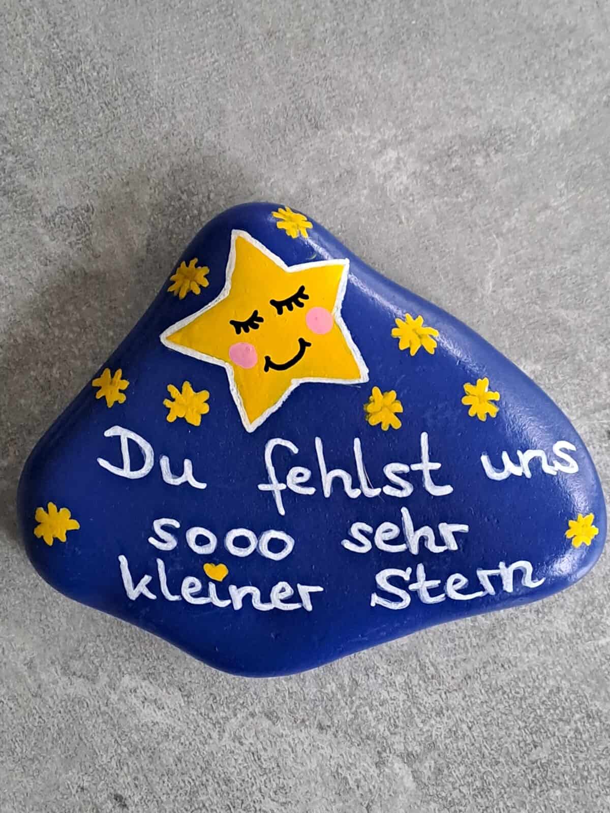 Dunkelblau lackierter Stein mit einem großen gelben Stern mit schlafendem Gesicht darauf, daneben viele kleine gelbe Steine und der Spruch Du fehlst uns sooo sehr kleiner Stern, Variante 2