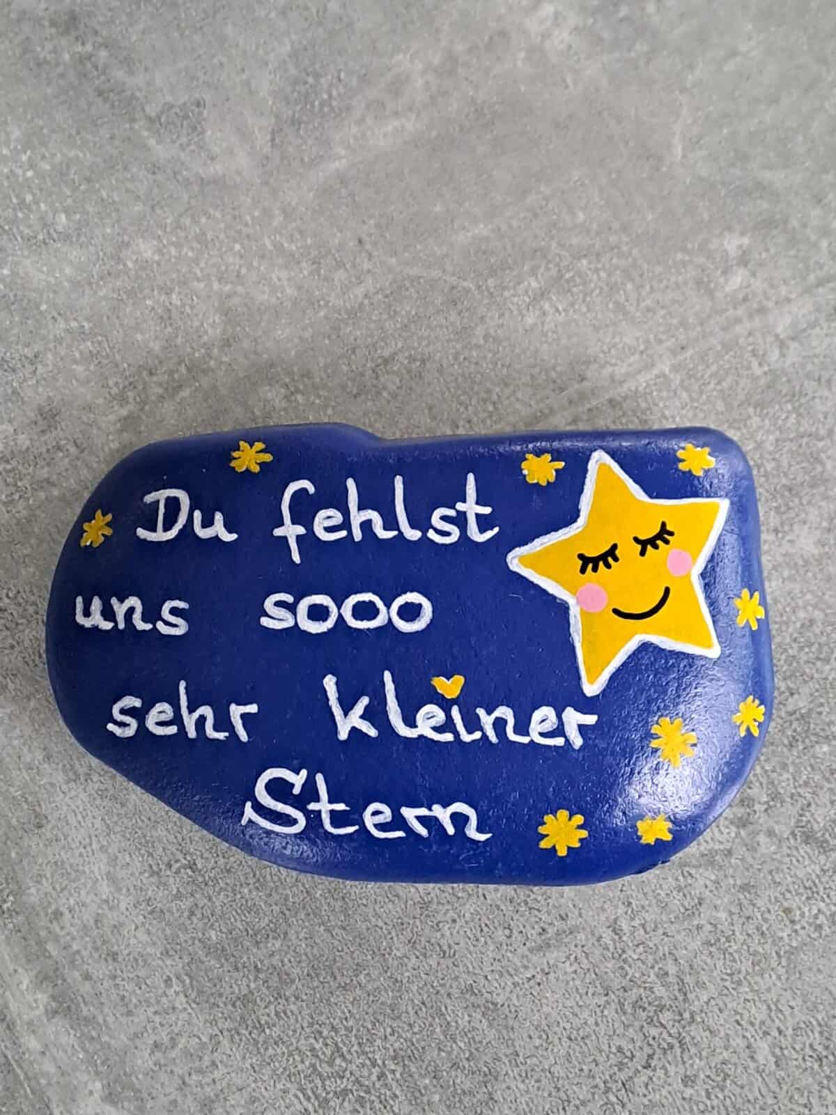Dunkelblau lackierter Stein mit einem großen gelben Stern mit schlafendem Gesicht darauf, daneben viele kleine gelbe Steine und der Spruch Du fehlst uns sooo sehr kleiner Stern, Variante 3