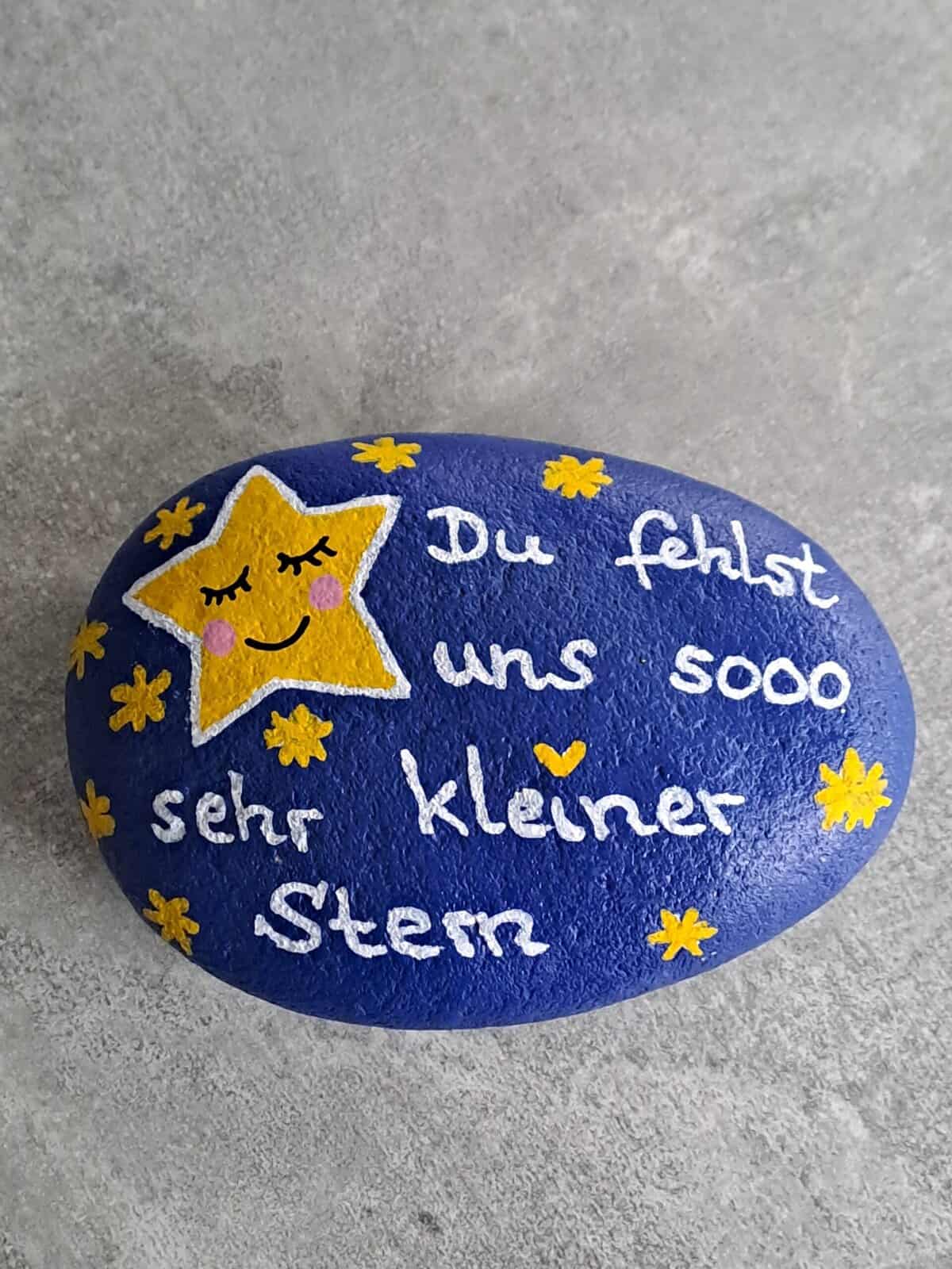 Dunkelblau lackierter Stein mit einem großen gelben Stern mit schlafendem Gesicht darauf, daneben viele kleine gelbe Steine und der Spruch Du fehlst uns sooo sehr kleiner Stern, Variante 4