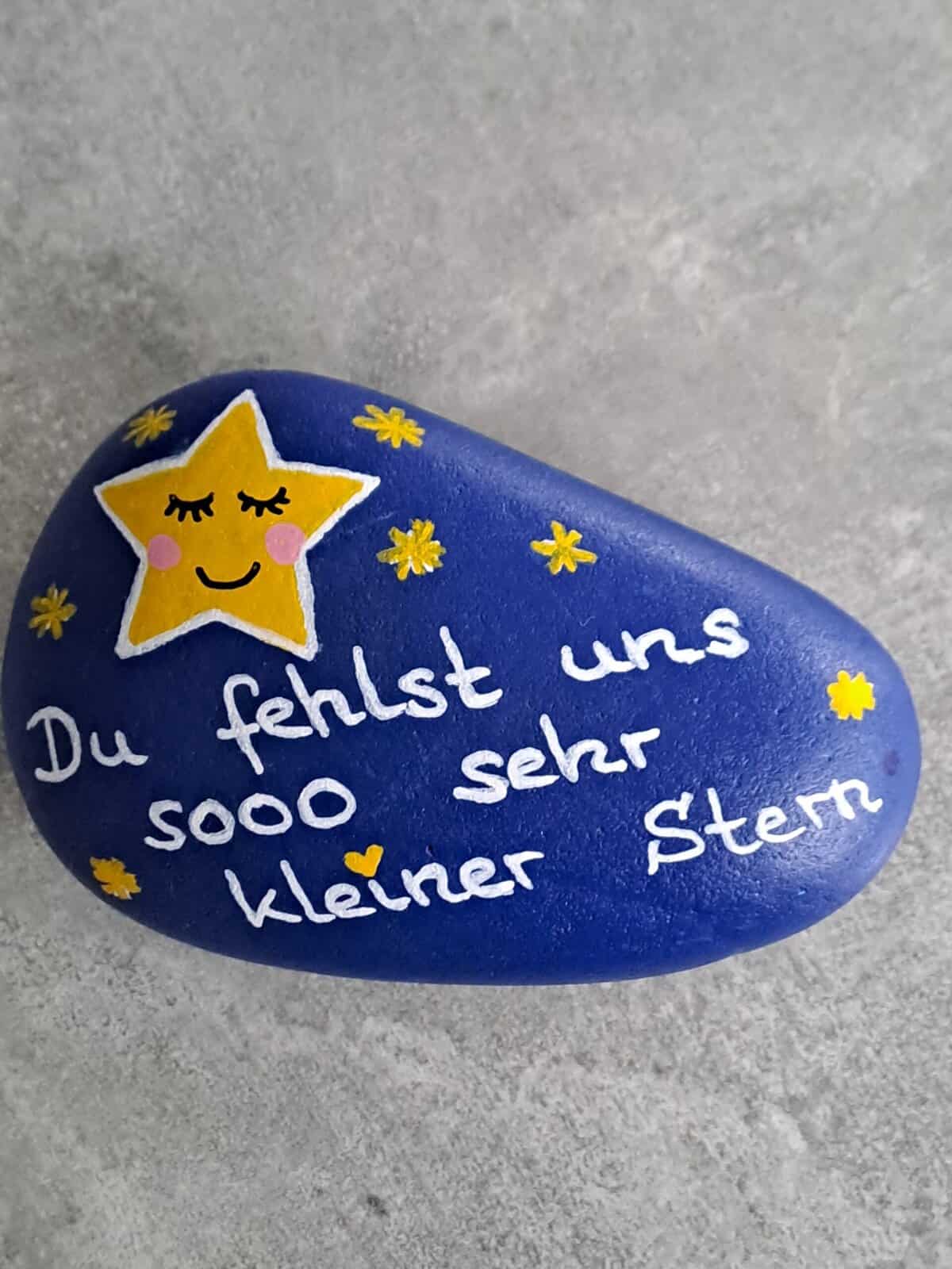 Dunkelblau lackierter Stein mit einem großen gelben Stern mit schlafendem Gesicht darauf, daneben viele kleine gelbe Steine und der Spruch Du fehlst uns sooo sehr kleiner Stern, Variante 5